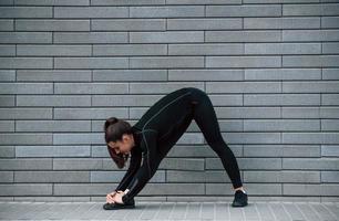 ung sportigt flicka i svart sportkläder utomhus håller på med stretching nära grå vägg foto