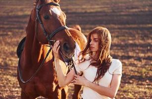 ung kvinna stående med henne häst i lantbruk fält på solig dagtid foto