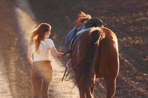 ung kvinna i skyddande hatt gående med henne häst i lantbruk fält på solig dagtid foto