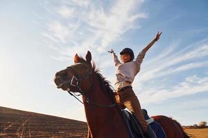 ung kvinna i skyddande hatt med henne häst i lantbruk fält på solig dagtid foto