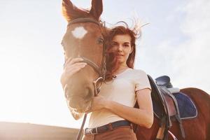 skön solsken. ung kvinna stående med henne häst i lantbruk fält på dagtid foto