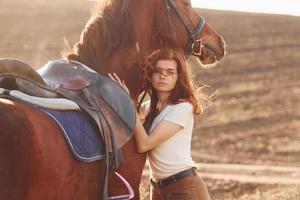 ung kvinna fattande henne häst i lantbruk fält på solig dagtid foto