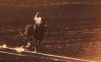 ung kvinna i skyddande hatt ridning henne häst i lantbruk fält på solig dagtid foto