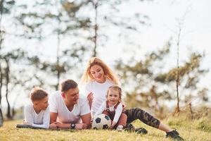 Sammanträde och liggande på de jord. Lycklig familj utgifterna helgen tillsammans utomhus nära de skog. med dotter och son foto