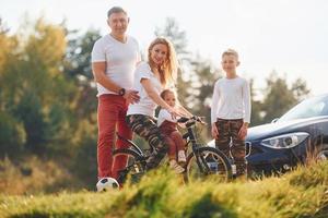 med Cyklar nära de bil. Lycklig familj utgifterna helgen tillsammans utomhus nära de skog. med dotter och son foto