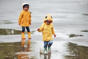 två barn i gul vattentät kappor och stövlar spelar utomhus efter de regn tillsammans foto