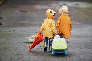 två barn med paraply, resväska och gul vattentät kappor och stövlar gående utomhus efter de regn tillsammans foto