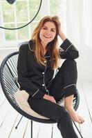 glad ung kvinna i svart pyjamas Sammanträde på stol inomhus på dagtid foto