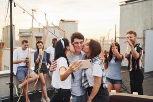 två flickor kissing ett man. tar selfie. grupp av ung människor i tillfällig kläder ha en fest på taket tillsammans på dagtid foto