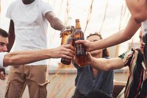 håller på med Skål förbi flaskor av öl. grupp av ung människor i tillfällig kläder ha en fest på taket tillsammans på dagtid foto