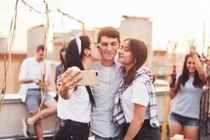 två flickor kissing ett man. tar selfie. grupp av ung människor i tillfällig kläder ha en fest på taket tillsammans på dagtid foto