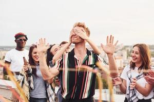 mannens ögon täckt förbi händer. grupp av ung människor i tillfällig kläder ha en fest på taket tillsammans på dagtid foto