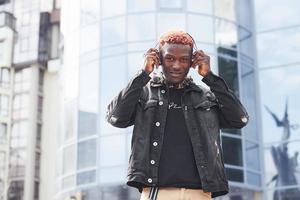lyssnande till musik i hörlurar. ung afrikansk amerikan man i svart jacka utomhus i de stad stående mot modern företag byggnad foto
