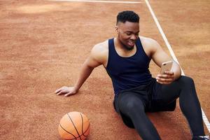 använder sig av telefon. afrikansk amerikan man spelar basketboll på de domstol utomhus foto