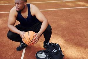 sitter med svart väska och framställning för de spel. afrikansk amerikan man spelar basketboll på de domstol utomhus foto