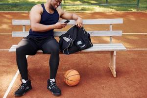 sitter med svart väska och framställning för de spel. afrikansk amerikan man spelar basketboll på de domstol utomhus foto
