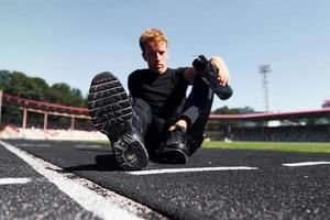 trött löpare sitter på Spår och tar en ha sönder. sportigt ung kille i svart skjorta och byxor utomhus på dagtid foto