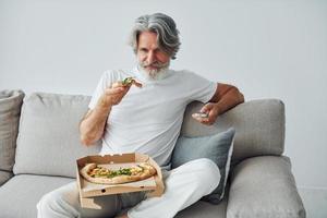 äter utsökt pizza medan tittar på TV show. senior eleganta modern man med grå hår och skägg inomhus foto