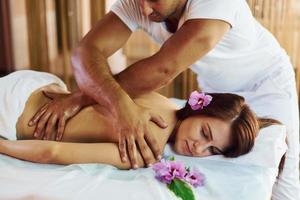 lila blomma liggande ner. man gör massage till de ung kvinna i vit handduk inomhus foto