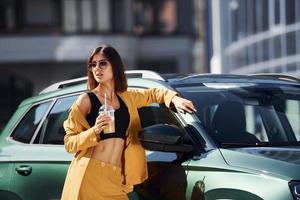 med dryck i händer. ung modern kvinna i vinröd färgad täcka på dagtid med henne bil foto
