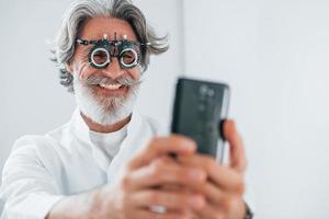 senior man med grå hår och skägg är i oftalmologi klinik ler och innehar smartphone foto