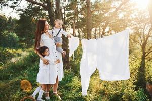 vit kläder hängande på de rep till torr. ung mor med henne liten dotter och son är utomhus i de skog. skön solsken foto