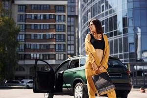 modern företag byggnad på bakgrund. ung modern kvinna i vinröd färgad täcka på dagtid med henne bil foto