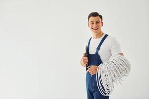 innehar kablar, anteckningsblock och skruvmejsel. manlig arbetstagare i blå enhetlig stående inuti av studio mot vit bakgrund foto