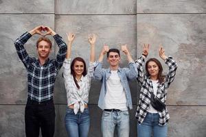 framställning annorlunda gester. grupp av ung positiv vänner i tillfällig kläder stående tillsammans mot grå vägg foto
