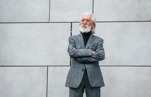 senior affärsman i formell kläder, med grå hår och skägg är utomhus står mot de vägg foto