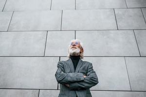 senior affärsman i formell kläder, med grå hår och skägg är utomhus står mot de vägg foto