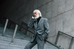 senior affärsman i formell kläder, med grå hår och skägg är utomhus samtal förbi telefon foto