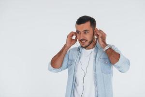 lyssnar till de musik i hörlurar. ung stilig man stående inomhus mot vit bakgrund foto