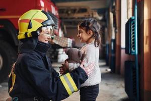 Lycklig liten flicka är med kvinna brandman i skyddande enhetlig foto