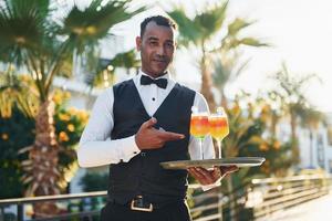 innehar cocktails. svart servitör i formell kläder är på hans arbete utomhus på solig dagtid foto