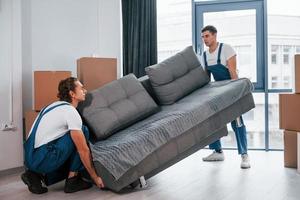 bärande tung soffa. två ung flyttare i blå enhetlig arbetssätt inomhus i de rum foto