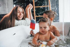 ung mor hjälper henne son och dotter. två barn tvättning i de bad foto