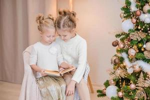 2 blond flickor är Sammanträde på ett fåtölj förbi de jul träd och läsning en bok. mysigt kvällar med en bok förbi de träd. flick läsa en bok tillsammans. värma relation foto