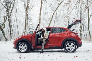 ny bil är i de skog. skön ung kvinna är utomhus nära henne röd bil på vinter- tid foto