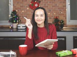 asiatisk kvinna bär röd stickat Tröja Sammanträde på tabell med röd kopp av kaffe och gåva lådor i de kök med jul dekoration, använder sig av penna skrivning namn lista på anteckningsbok. foto