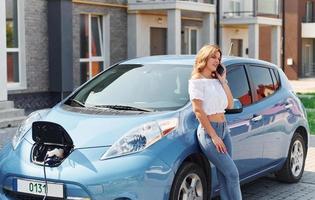 stående nära de bil. ung kvinna i tillfällig kläder med henne elektromobil utomhus på dagtid foto
