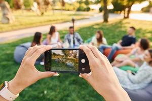 framställning Foto förbi använder sig av smartphone. grupp av ung människor ha en fest i de parkera på sommar dagtid