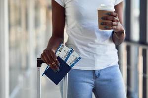 ung afrikansk amerikan kvinna passagerare i tillfällig kläder är i flygplats med biljetter och kopp av dryck foto