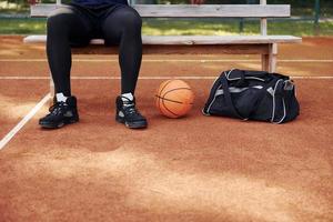sitter med svart väska och framställning för de spel. afrikansk amerikan man spelar basketboll på de domstol utomhus foto