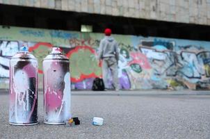 flera Begagnade spray burkar med rosa och vit måla lögn på de asfalt mot de stående kille i främre av en målad vägg i färgad graffiti ritningar foto