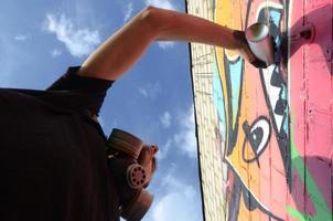 ung graffiti konstnär med ryggsäck och gas mask på hans nacke målarfärger färgrik graffiti i rosa toner på tegel vägg. gata konst och samtida målning bearbeta foto