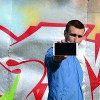 de graffiti konstnär demonstrerar en smartphone med ett tömma svart skärm mot de bakgrund av en färgrik målad vägg. gata konst begrepp foto