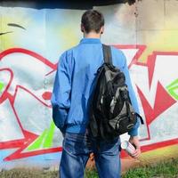 en ung graffiti konstnär med en svart väska utseende på de vägg med hans graffiti på en vägg. gata konst begrepp foto