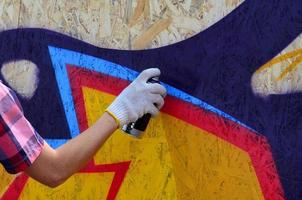 en hand med en spray kan den där drar en ny graffiti på de vägg. Foto av de bearbeta av teckning en graffiti på en trä- vägg närbild. de begrepp av gata konst och olaglig vandalism