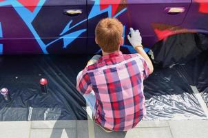 en ung rödhårig graffiti konstnär målarfärger en ny färgrik graffiti på de bil. Foto av de bearbeta av teckning en graffiti på en bil närbild. de begrepp av gata konst och olaglig vandalism